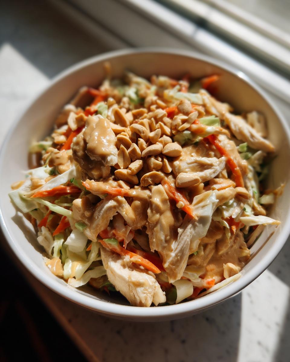 Close-up of a bowl filled with vibrant thai chicken salad, shredded cabbage, carrots, chicken, and topped with peanut dressing and chopped peanuts.