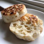 Close-up of a homemade english muffin, split open to show the airy interior and toasted golden-brown surfaces.