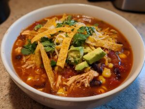 Close-up of a white bowl filled with rich, red tortilla soup, topped with shredded chicken, avocado, cheese, and tortilla strips.
