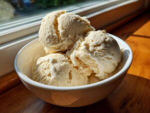 Three scoops of creamy vanilla protein ice cream melting slightly in a white bowl near a sunlit window.