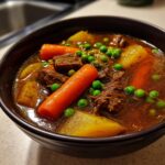 Close-up of a rich, dark broth vegetable beef soup featuring chunks of beef, bright orange carrots, potatoes, and green peas.
