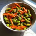 Close-up of a bright green edamame salad mixed with sliced carrots and topped with sesame seeds.