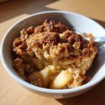 Close-up of a warm serving of apple crumble with a golden brown topping in a white bowl.