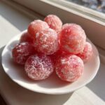 A pile of pink, round watermelon mochi dusted heavily with white sugar, sitting on a small white plate.