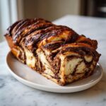 A freshly baked, whole chocolate babka loaf showcasing rich, dark chocolate swirls and a glossy top, resting on a white plate.