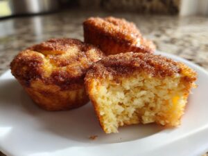 Three air fryer cinnamon sugar french toast muffins on a white plate, one is bitten to show the fluffy interior.