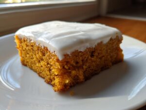 A close-up of a single, moist slice of orange pumpkin bars topped with thick white cream cheese frosting.