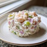 A mound of creamy seafood salad featuring shrimp, celery, and red onion, served on a white plate.