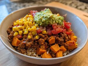 A close-up of an amazing sweet potato taco bowl layered with rice, seasoned meat, roasted sweet potatoes, corn, tomatoes, and guacamole.