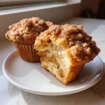 Two apple cinnamon muffins on a white plate, one broken open showing tender crumb and apple chunks.