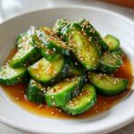 Close-up of vibrant green cucumber slices in a savory brown dressing, topped generously with toasted sesame seeds for an asian cucumber salad.