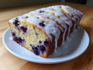 A loaf of moist blueberry zucchini bread topped with a thick white glaze, sitting on a white plate.