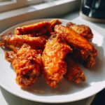 Close-up of several crispy, glossy buffalo chicken wings piled on a white plate.