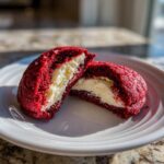 A bright red velvet cookie cut in half revealing a thick, creamy cheesecake filling, perfect for cheesecake stuffed red velvet cookies.