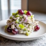 A mound of creamy chicken protein salad mixed with celery, red onion, and topped with red grapes on a white plate.