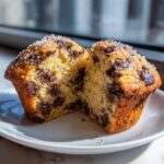 A delicious chocolate chip muffin cut in half, showing the moist interior packed with chocolate chips and topped with coarse sugar.