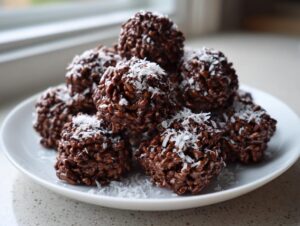 A pile of rich chocolate rice krispie balls with coconut sprinkled on top, served on a white plate.