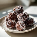A stack of rich chocolate rice krispie balls coated in shredded coconut on a white plate.