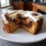 A cross-section view of a moist cinnamon roll cake showing the swirl filling and thick cream cheese frosting on top.