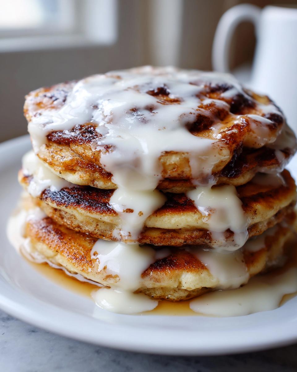 A tall stack of three golden cinnamon roll pancakes generously drizzled with white cream cheese icing.