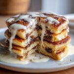 Close-up of a stack of fluffy cinnamon roll pancakes cut open, dripping with white cream cheese icing.