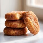 A stack of three soft churro cookies generously coated in cinnamon sugar, with one cookie leaning against the stack.