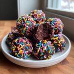 A pile of chocolate cosmic brownie protein bites covered in colorful rainbow sprinkles on a white plate.