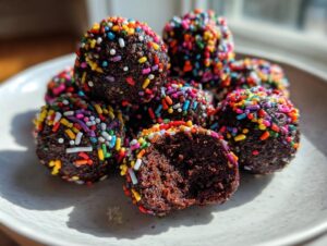 A pile of dark chocolate cosmic brownie protein bites covered in rainbow sprinkles, one is cut in half.
