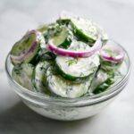 Close-up of sliced cucumbers coated in a creamy dressing, topped with red onion rings and fresh dill, showcasing the creamy cucumber salad.
