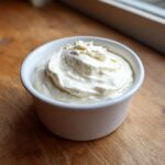 A small white bowl filled with thick, creamy homemade garlic aioli recipe, set on a wooden surface.