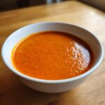 Close-up of a white bowl filled with vibrant, creamy roasted red pepper soup on a wooden table.