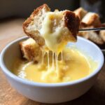 A piece of crusty bread being dipped into a bowl of melted cheese fondue recipe, showing long cheese pulls.