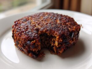 A close-up of a perfectly cooked, dark brown easy black bean burger patty with a bite taken out, resting on a white plate.