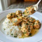 A spoonful of tender chicken scampi pieces coated in garlic butter sauce being lifted over a bed of garlic parmesan rice.