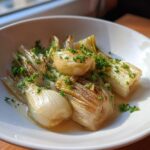 Close-up of tender french braised leeks served in a white bowl with broth and fresh parsley.