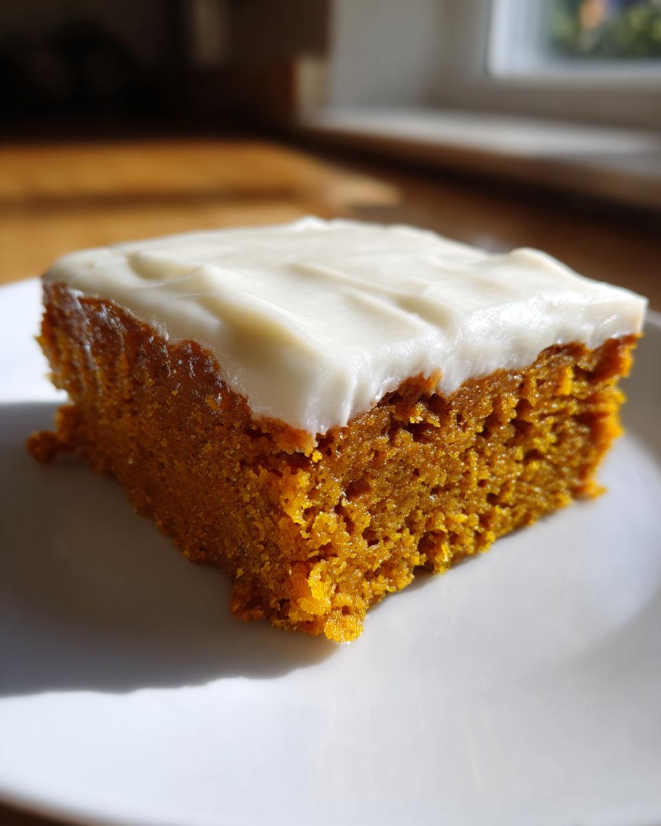 A moist, square slice of orange pumpkin bars topped with thick cream cheese frosting on a white plate.