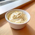 A small white bowl filled with creamy, pale yellow garlic aioli recipe without mayo, topped with dried herbs.
