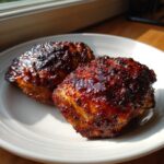 Two perfectly cooked, glazed bbq chicken thighs with a dark, caramelized crust served on a white plate.