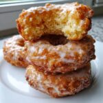 Close-up of a stack of three freshly glazed cake donuts, with the top one having a bite taken out showing the crumb.