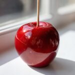 Close-up of a single, perfectly glossy, deep red candy apple resting on a white surface near a window.