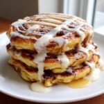 A tall stack of three golden gluten free cinnamon roll pancakes drizzled heavily with white cream cheese icing.