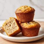 A stack of three golden brown protein muffins, one cut in half showing the moist interior.