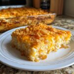 A close-up of a square serving of creamy hashbrown casserole topped with melted cheese and crispy corn flakes.