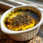 Close-up of a white ramekin filled with olive oil, balsamic vinegar, and a generous topping of herbs and spices for balsamic and olive oil bread dip.