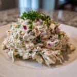 A mound of creamy high protein chicken salad mixed with red onion and topped with fresh parsley on a white plate.