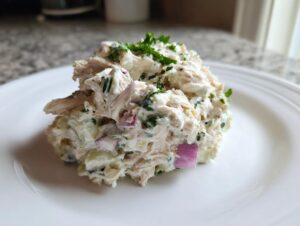 A scoop of creamy high protein chicken salad mixed with red onion and herbs, served on a white plate.