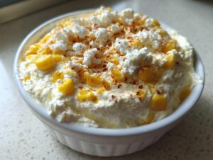 Close-up of creamy, white high protein elote dip topped with yellow corn kernels, crumbled white cheese, and paprika.