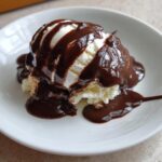 A scoop of vanilla ice cream generously covered in glossy, homemade chocolate sauce served in a white bowl.