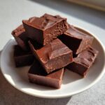 A stack of rich, dark chocolate squares of keto chocolate peanut butter fudge piled on a small white plate.