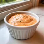 Close-up of creamy, orange-hued low carb big mac sauce speckled with herbs in a white fluted dipping bowl.